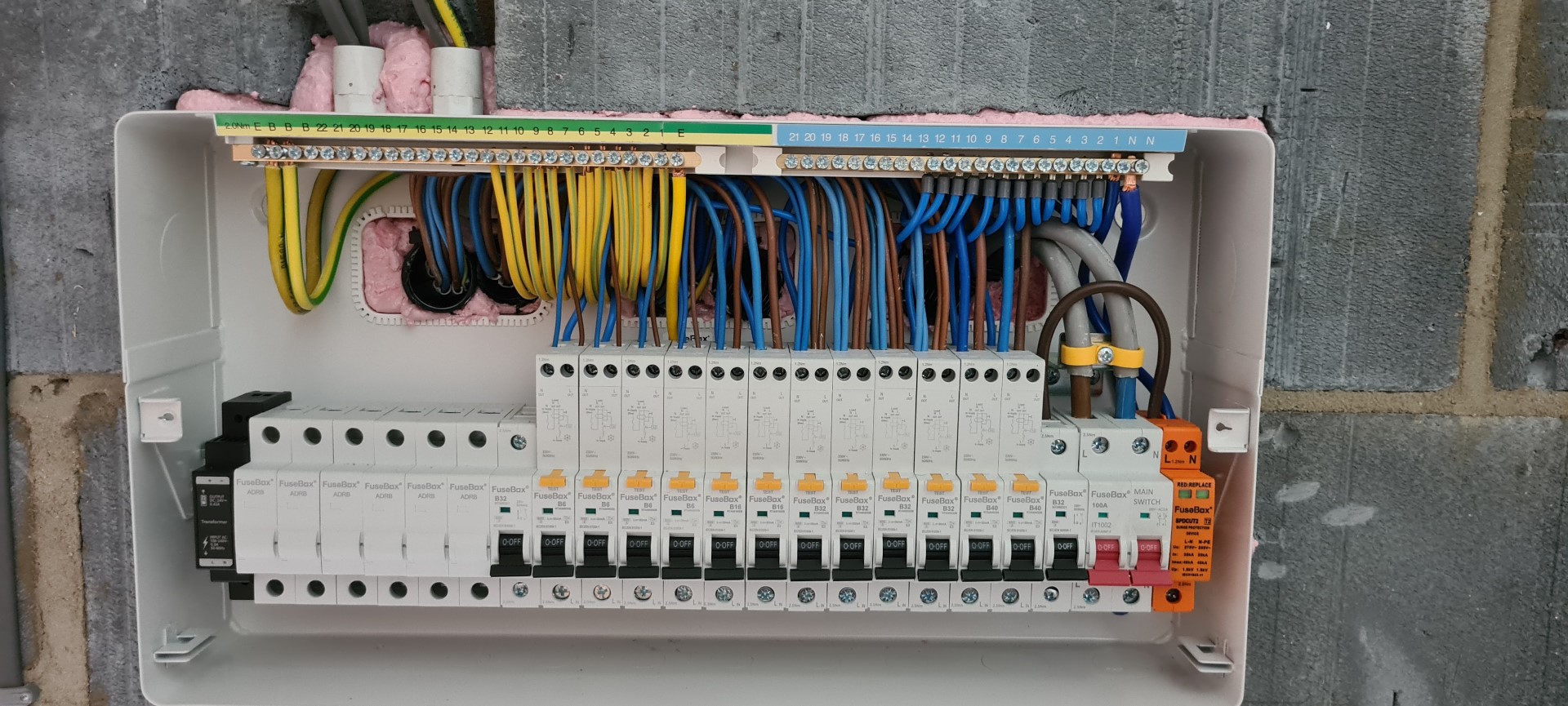 A detailed image of an open electrical distribution board installed by H Noble & Sons LTD, showcasing an array of neatly arranged cables in pink, yellow, and blue, connected to circuit breakers and an earth terminal block. The board is mounted on a concrete wall with visible insulation, reflecting the meticulous workmanship typical of H Noble & Sons LTD's electrical services