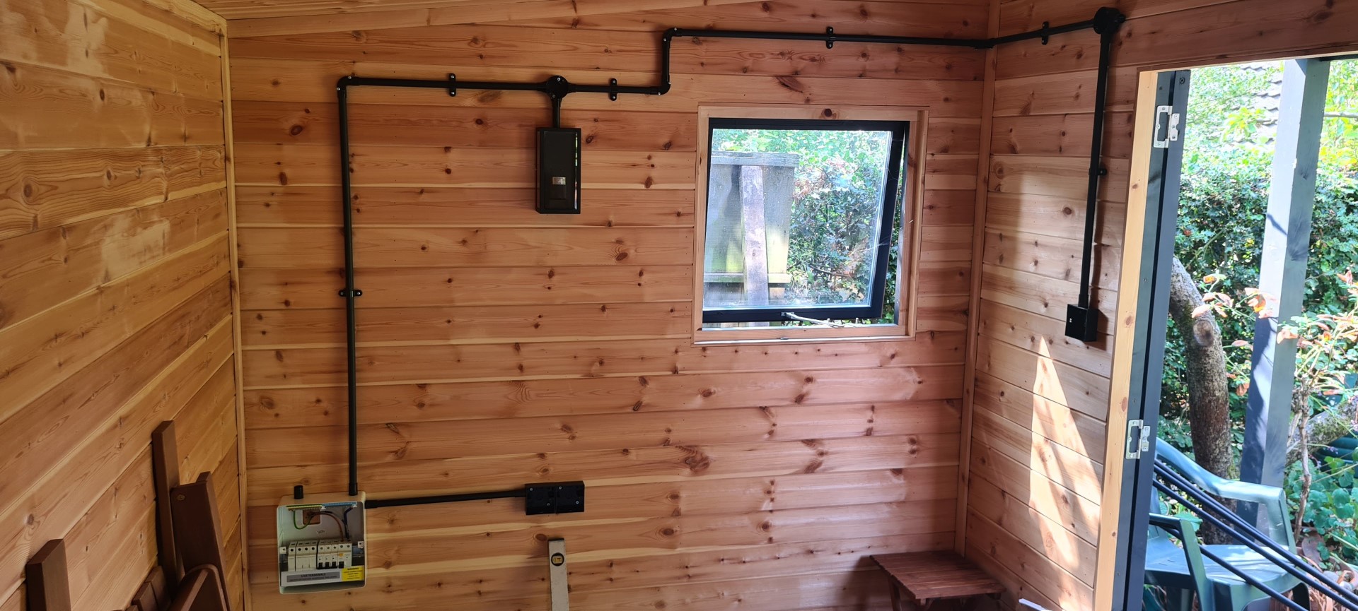 An indoor view of a cabin with H Noble & Sons LTD electrical installation. The walls are lined with natural pine cladding and feature black electrical conduits. A small window framed by the wooden interior looks out to lush greenery, indicating a tranquil setting. The electrical setup includes a prominently displayed fuse box and other devices, showcasing the company's expertise in creating functional spaces